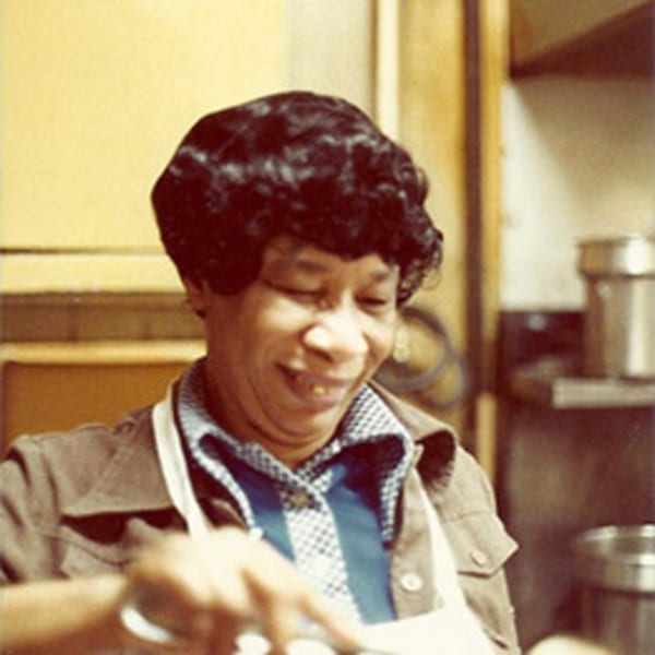 A smiling woman with dark curly hair and a gold tooth, wearing an apron, working in a diner kitchen.
