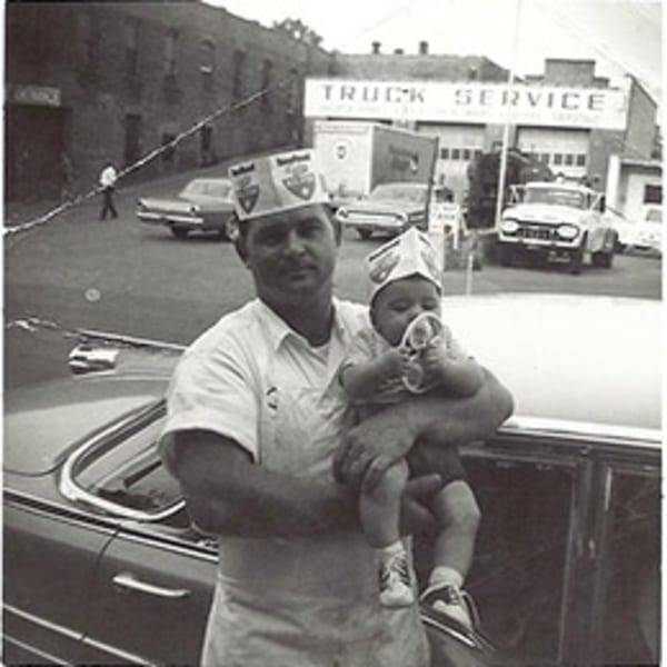 A man in a paper hat holds a baby, also in a paper hat, next to a car.