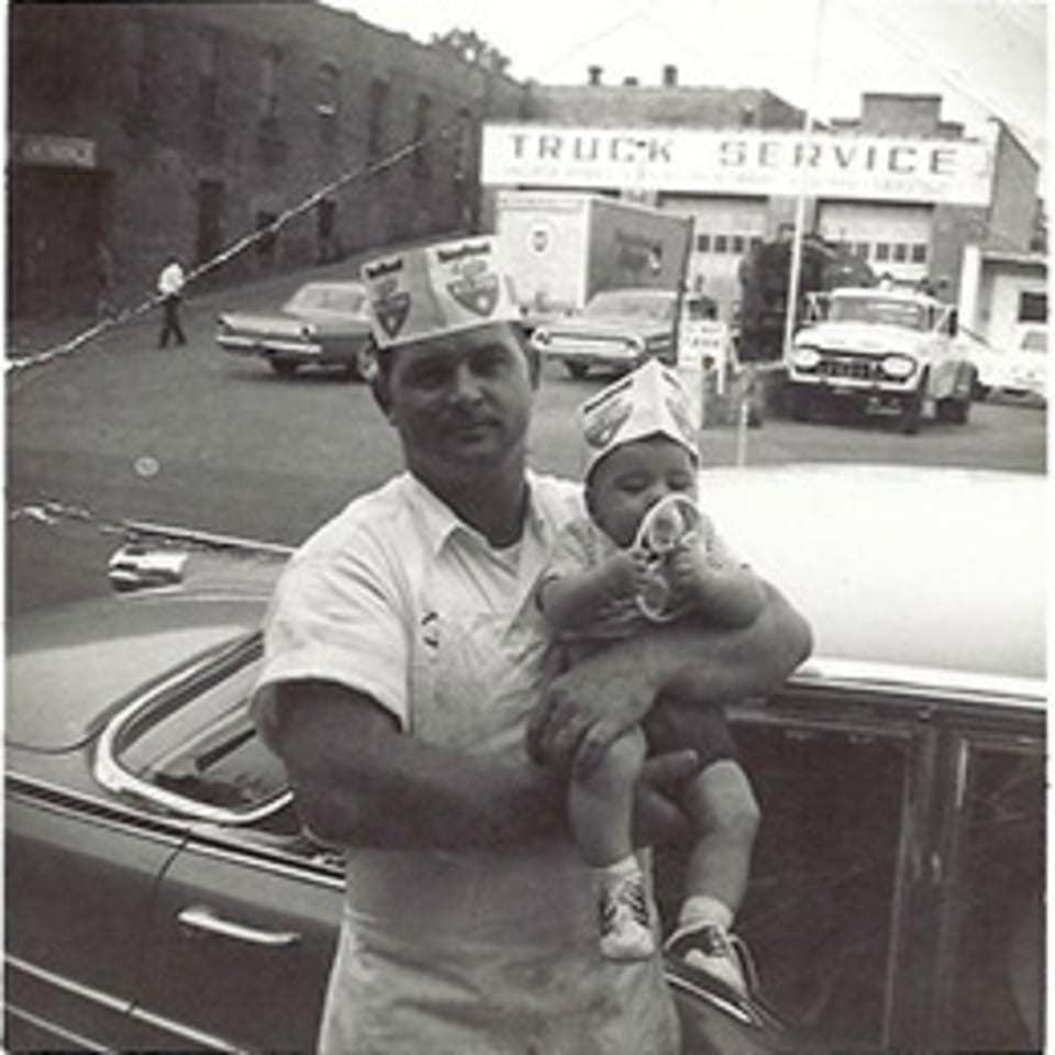 A man in a paper hat holds a baby, also in a paper hat, next to a car.