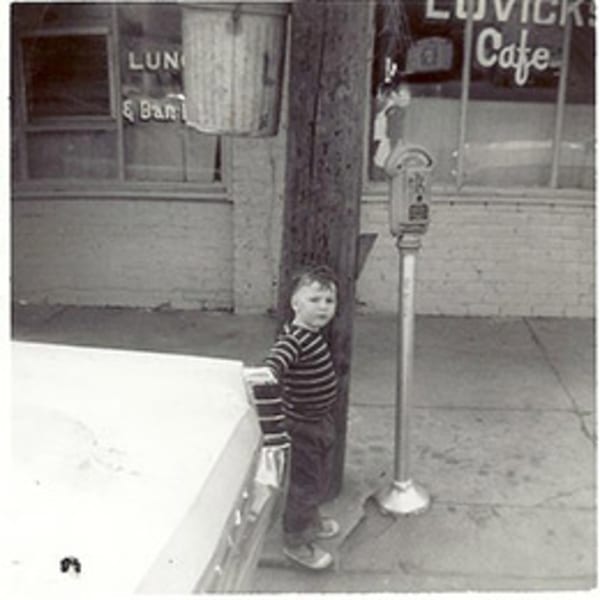 A young boy in a striped shirt stands beside a car and parking meter outside Lovick's Cafe.