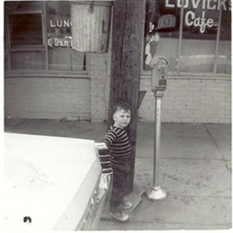 A young boy in a striped shirt stands beside a car and parking meter outside Lovick's Cafe.