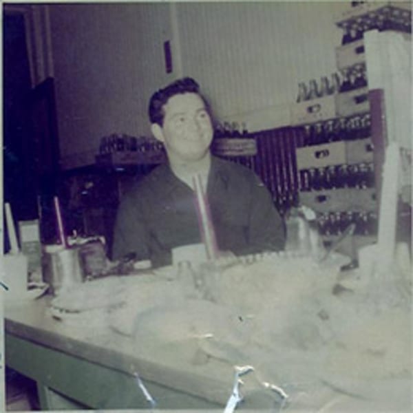 A man in a dark shirt smiles behind a diner counter with bottles stacked in the background.