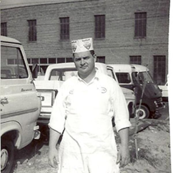 Black and white photo of a man in a chef's hat and apron standing in front of delivery vans.