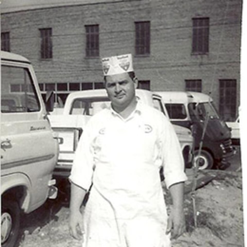 Black and white photo of a man in a chef's hat and apron standing in front of delivery vans.
