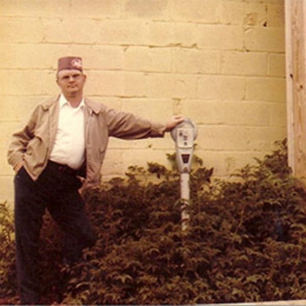 Man in a tan jacket and hat, leaning on a parking meter next to a yellow brick wall.