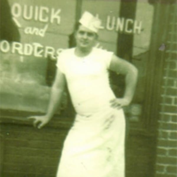A man in a white uniform and paper hat poses in front of a diner window.