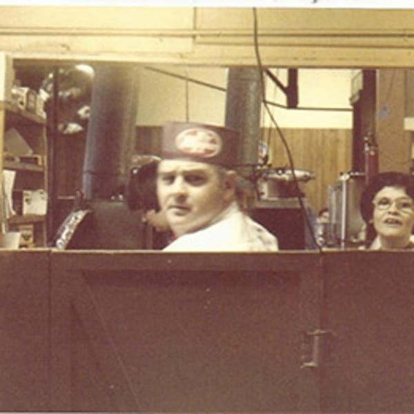 A man in a diner hat and a woman in glasses look over a counter in a vintage photo.