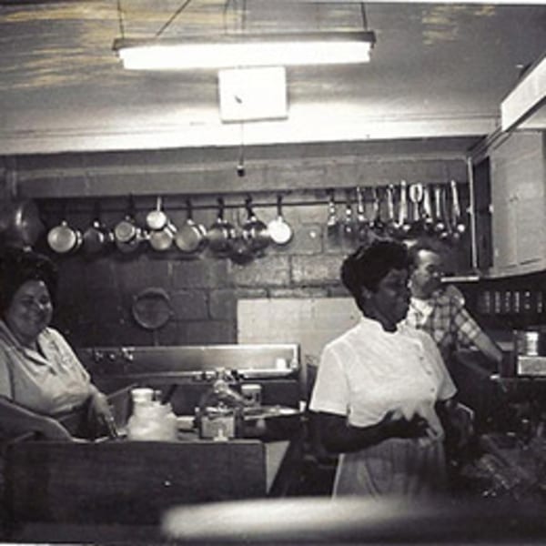 Black and white photo of three people working in a busy diner kitchen with hanging pots.