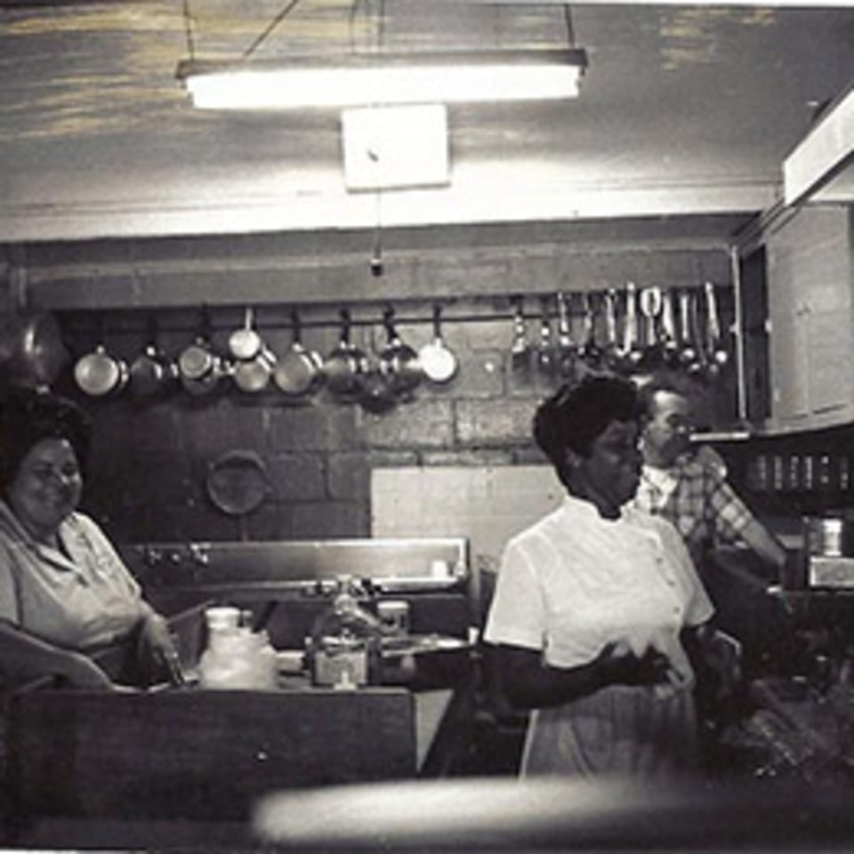Black and white photo of three people working in a busy diner kitchen with hanging pots.