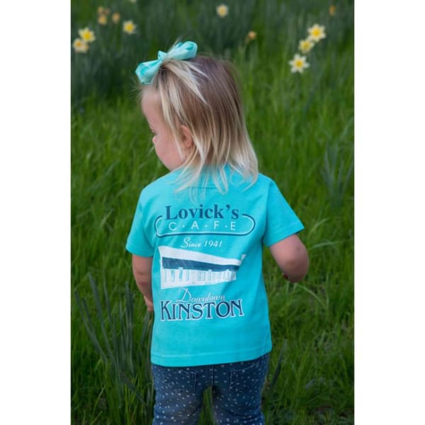A child from behind, wearing a Lovick's Cafe t-shirt, standing in a grassy field with flowers.