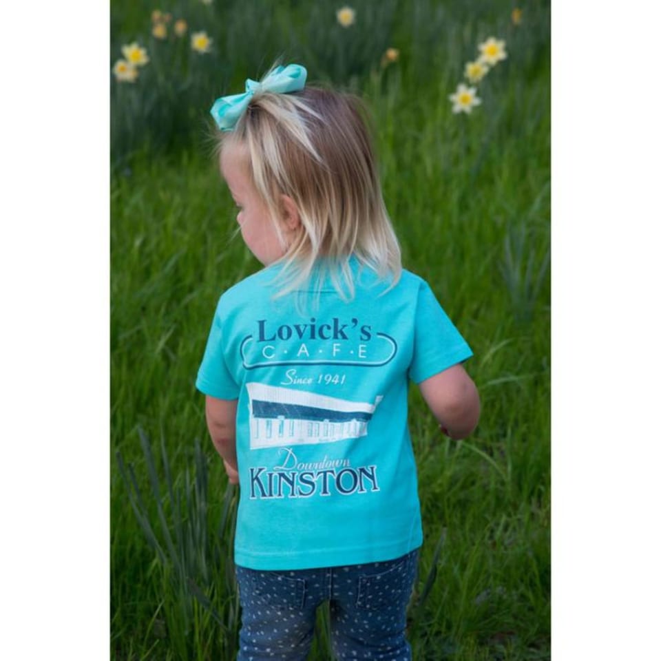 A child from behind, wearing a Lovick's Cafe t-shirt, standing in a grassy field with flowers.