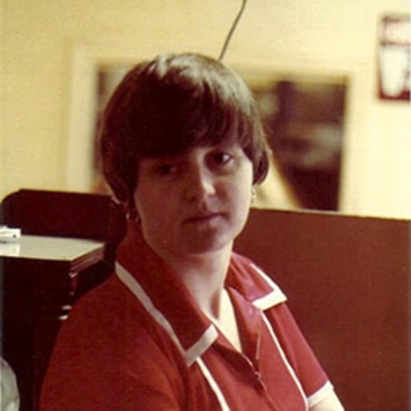 A woman with dark hair and a red and white collared shirt looks directly at the camera.