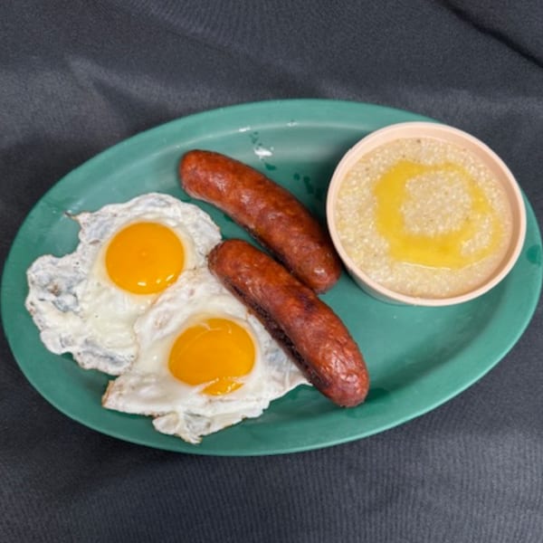 A diner breakfast of two fried eggs, two sausages, and a bowl of grits with butter on a green plate.