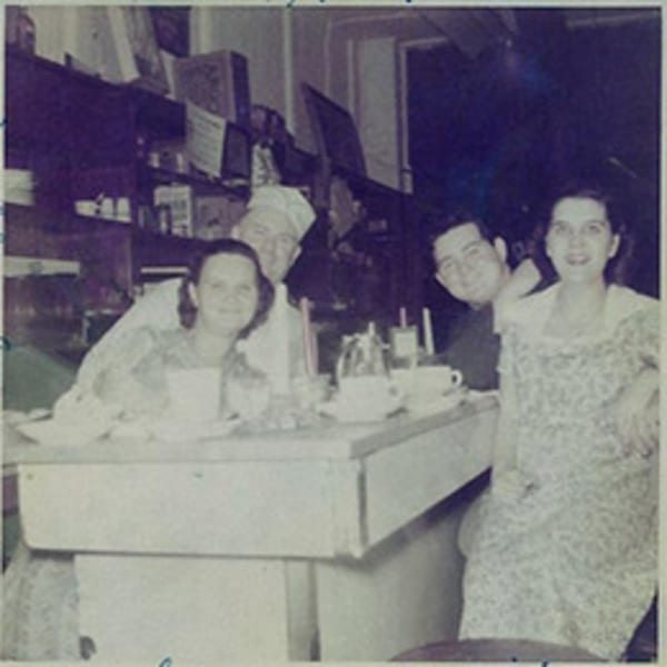 Four people, including a chef, smiling at a diner counter with coffee cups and a sugar dispenser.