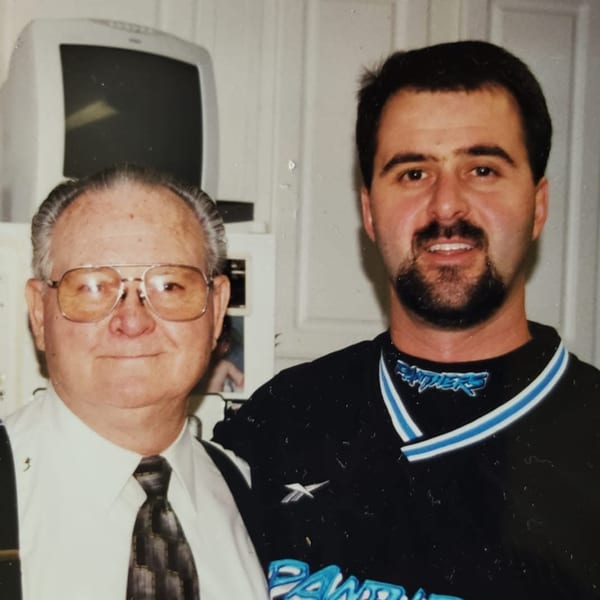 Two men, one older in a tie and glasses, one younger in a Panthers shirt, smiling.