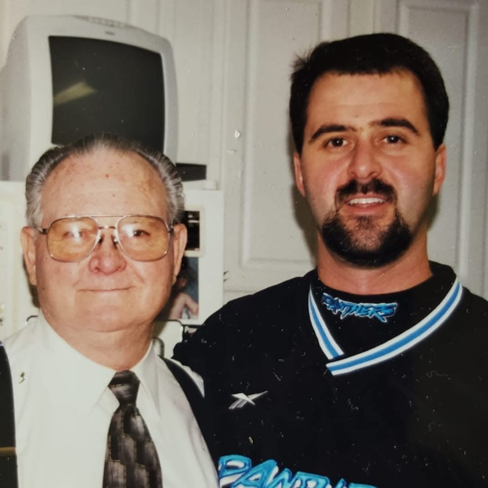 Two men, one older in a tie and glasses, one younger in a Panthers shirt, smiling.