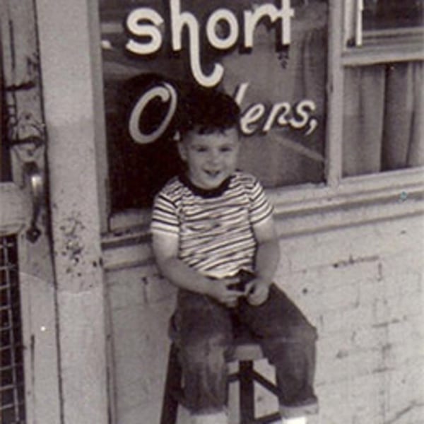 A young boy in a striped shirt sits on a stool outside Lovick's Cafe, smiling.