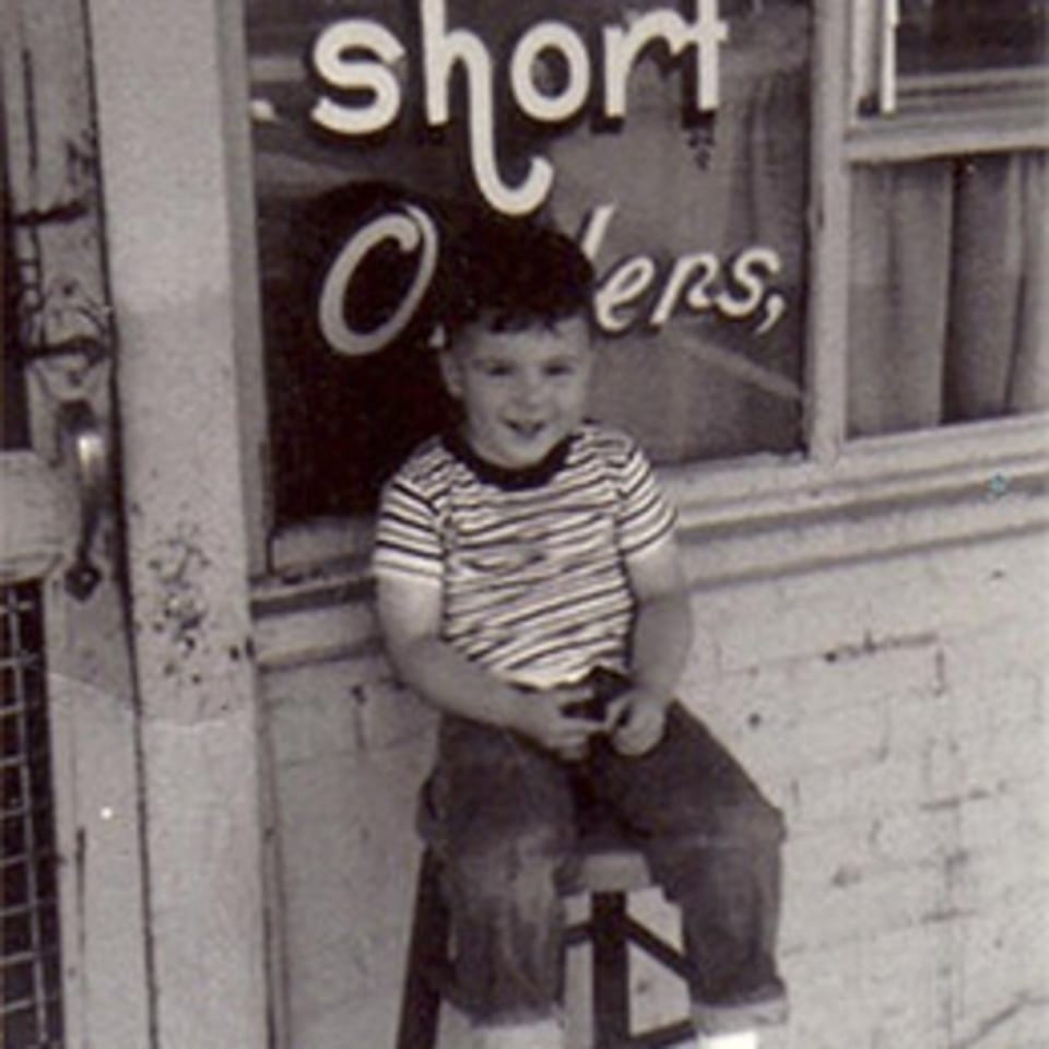 A young boy in a striped shirt sits on a stool outside Lovick's Cafe, smiling.