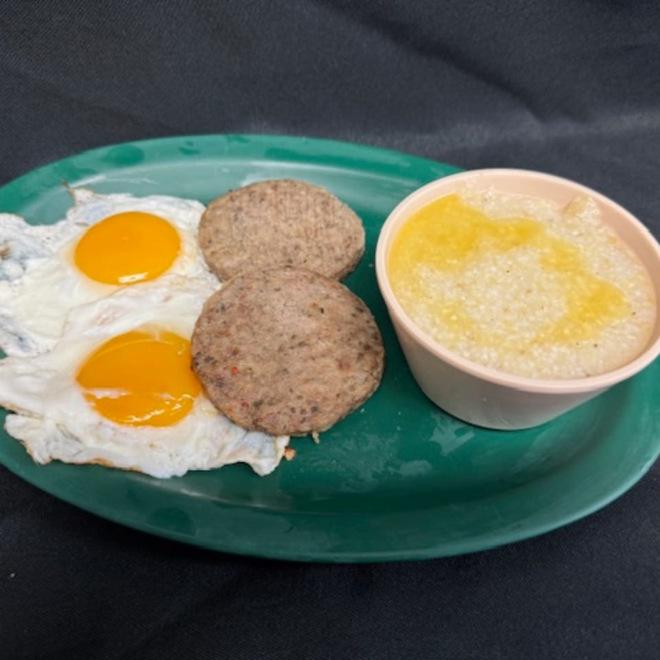 A diner breakfast on a green plate with two fried eggs, two sausage patties, and a cup of grits.