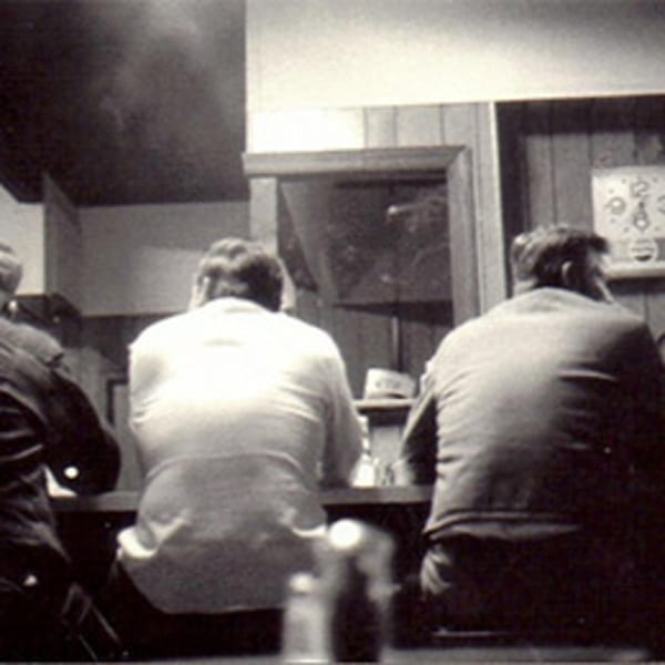 Three men seated at a counter, viewed from behind, in a vintage diner setting.